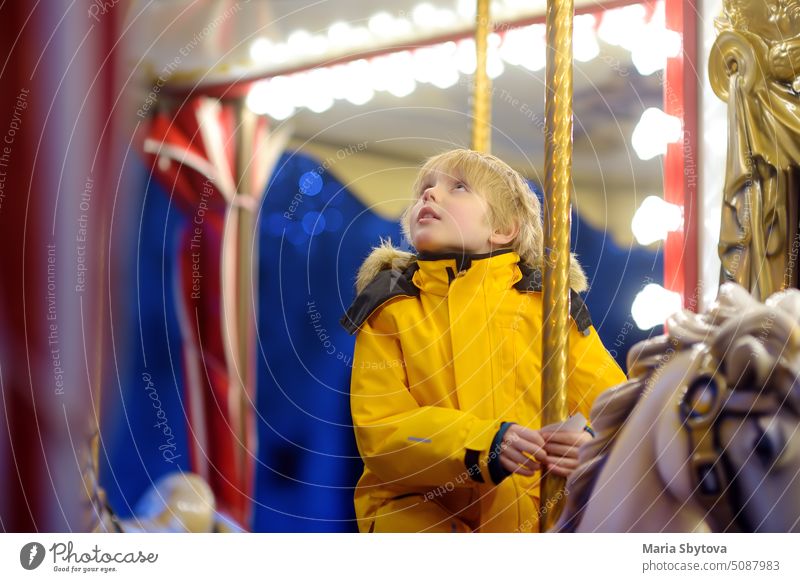 Cute blonde boy enjoying Christmas fair. Little child riding on a vintage carousel (merry go round). Outdoors entertainment activity for children on winter holidays