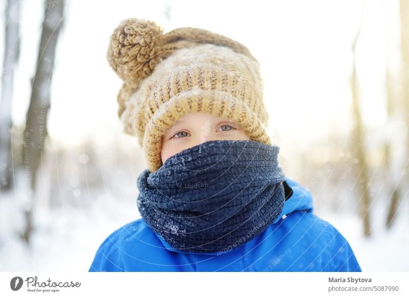 Portrait of little boy in blue winter clothes walks during snowfall on frost winter day. Child wearing warm clothing, hat, scarf is looking at you. Outdoors activities for kids in any weather