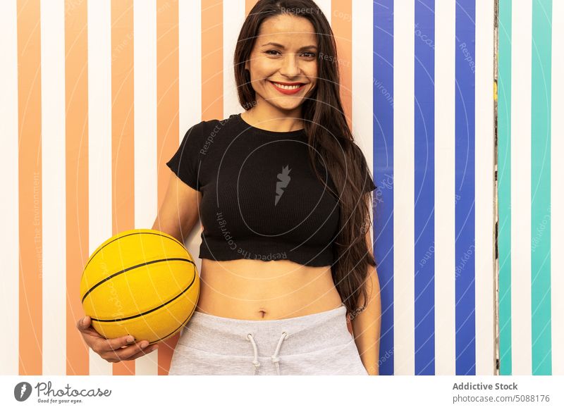 Smiling woman standing against striped wall with basketball ball sportswoman wellness hobby leisure physical player motivation sporty fit joy slim happy smile