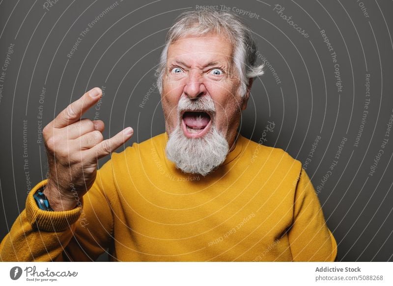 Senior man showing rock gesture portrait horn sign rock and roll cool senior elderly aged male gray hair beard yellow sweater pensioner studio shot retire