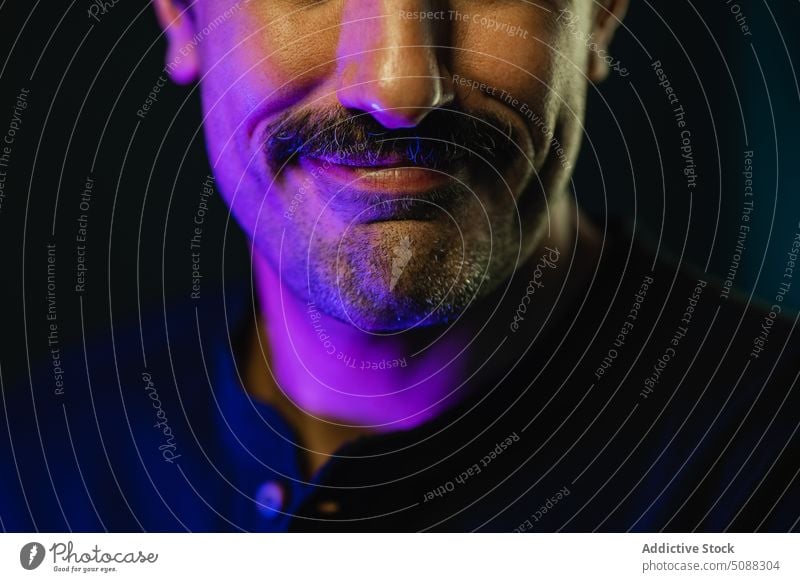 Crop face of man with mustache against black background glow neon light illuminate masculine studio shot darkness smile positive glad happy mood adult cheerful