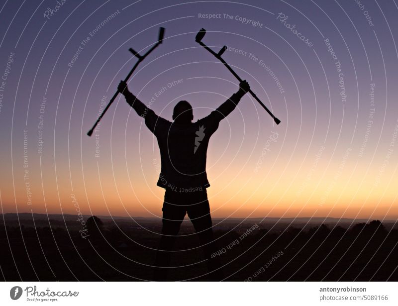Silhouette of man holding up crutches in celebration of reaching the top of a hill at sunset healed cured health healthcare patient pain freedom disability