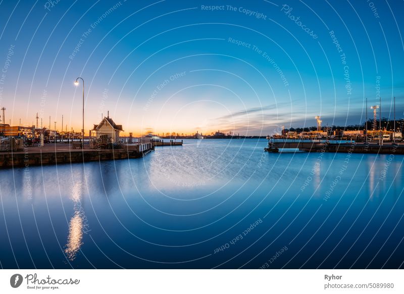 Helsinki, Finland. Landscape With City Pier, Jetty At Sunrise Time. Blue Sky Reflected In Tranquil Sea Water Surface. Berth In Lighting At Evening Night Illumination