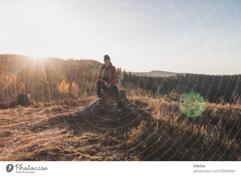 Active hiker sitting on a stump enjoys the feeling of reaching the top of the mountain at sunrise. A hiker is enlightened by the morning sun and enjoys the view. Beskydy mountains, Czech Republic