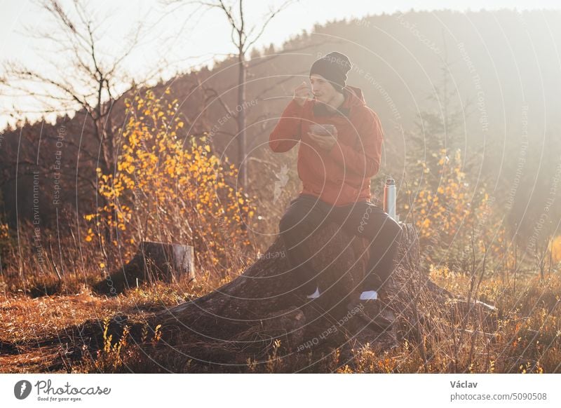 Tourist sitting on a stump eating porridge at sunrise. A hiker is enlightened by the morning sun and enjoys the view. Beskydy mountains, Czech Republic