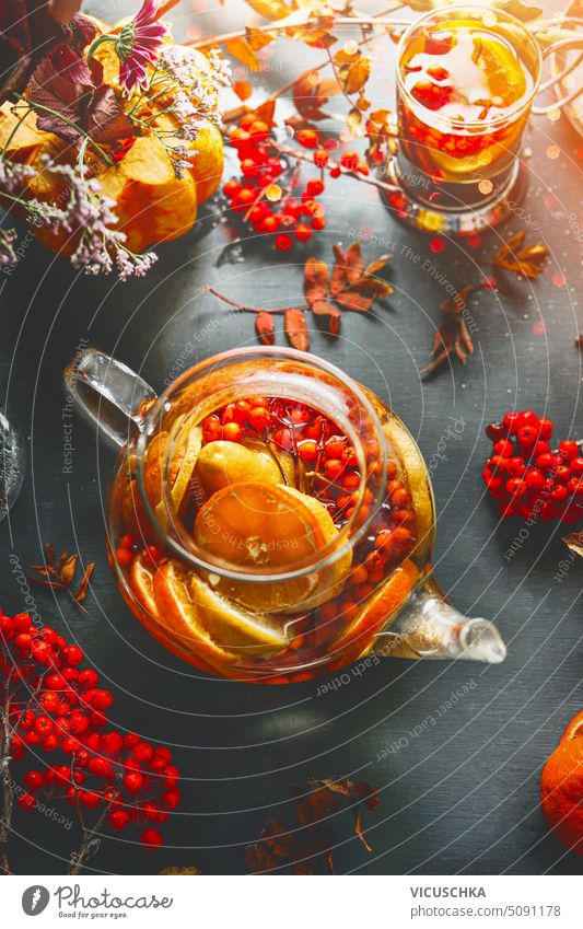 Class teapot with healthy vitamin tea with Rowan berries, orange and lemon slices on table with ingredients. Hot drink for cold autumn season. Close up class