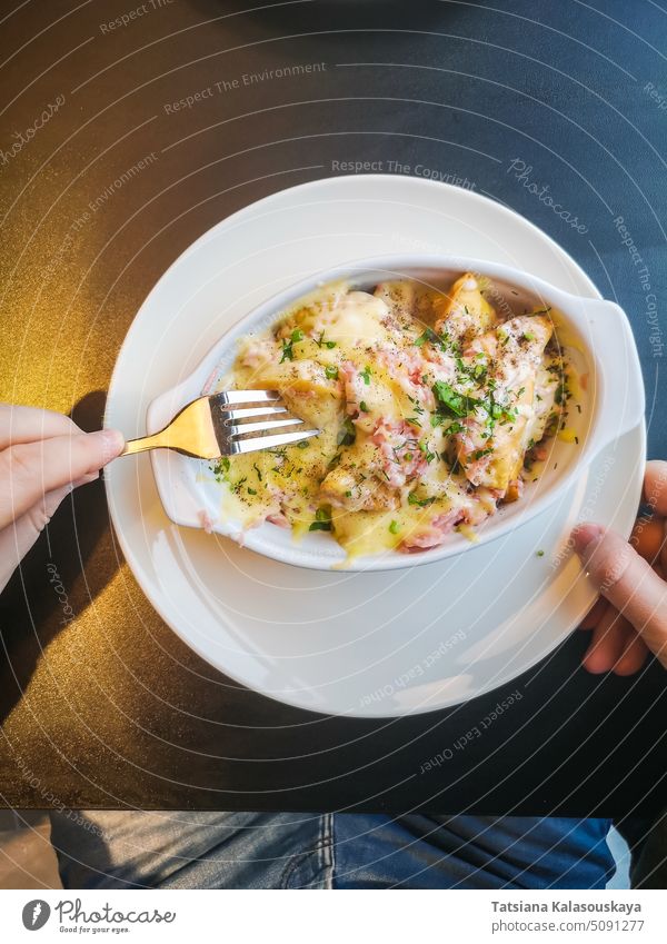 A man eats baked dumplings with a sauce of bacon, ham, herbs and cheese. hands Lithuanian national dish white plate black table food traditional lunch meal