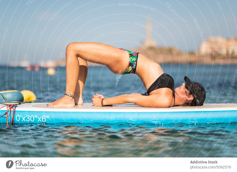Fit woman doing yoga asana on paddleboard in sea practice balance sup board swimwear healthy summer activity wellness fit wellbeing bridge pose