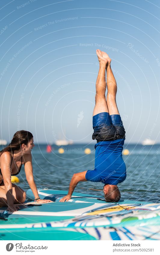 Man performing headstand in yoga while woman watching near sea paddleboard practice resort cloudless sirasana blue sky balance demonstrate nature aqua energy