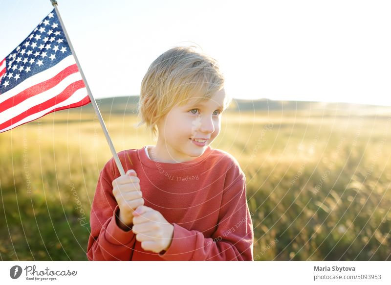 Cute little boy celebrating of July, 4 Independence Day of USA at sunny summer sunset. Child running and jumping with american flag symbol of United States over wheat field. Patriotism