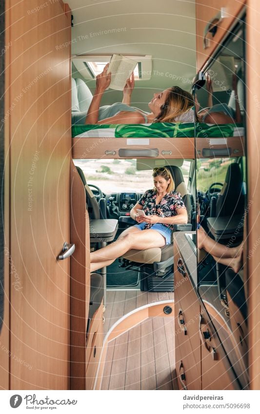 Woman lying on camper van bunk bed reading book while her friend looking cellphone woman female resting vehicle caravan bunkbed comfortable relaxation peace