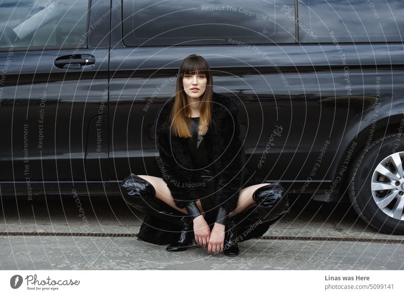 A gorgeous brunette model is posing for a model test. A fashionable woman dressed in black with some latex accents and a black car as a background. Hot wheels and even hotter beauty.