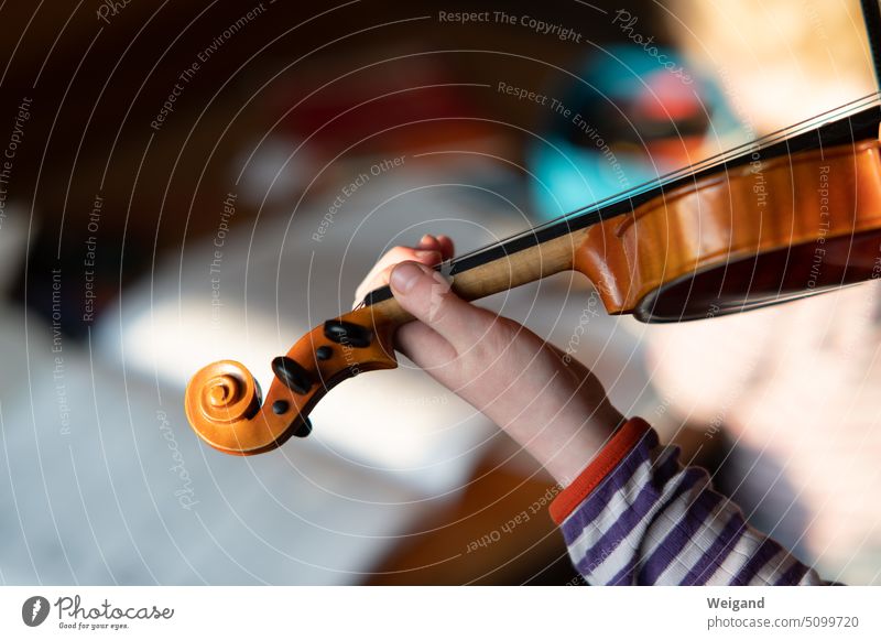 Close-up of a child's hand in the foreground holding and playing a violin, while a book of music can be glimpsed blurred in the background Child Violin Music