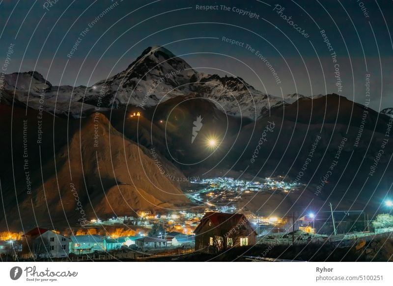 Stepantsminda, Georgia. Peak Of Mount Kazbek Covered With Snow, Famous Gergeti Church And Countryside Houses In Night Lightning view village town scenic europe