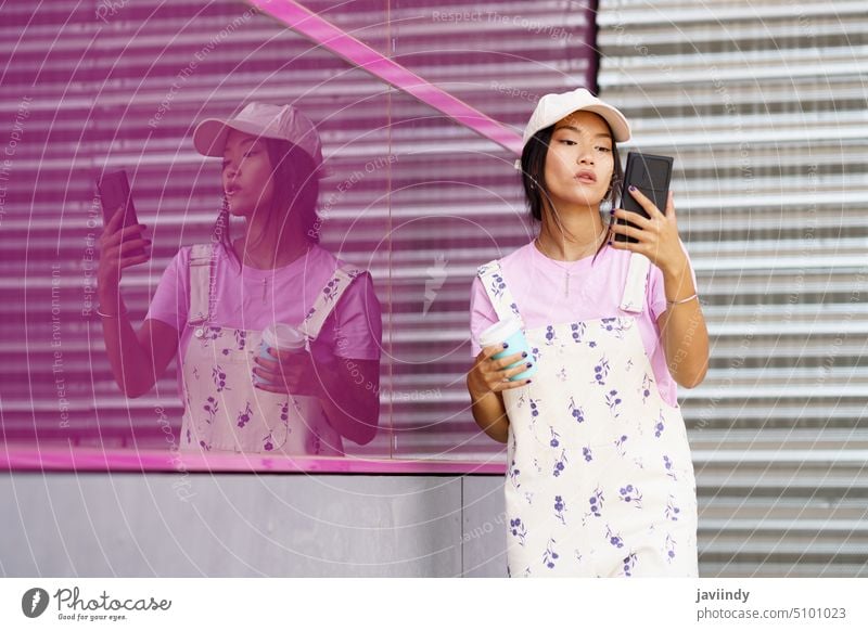 Young Asian woman with smartphone and coffee near pink wall using street read message style online building female social media glass wall reflection to go