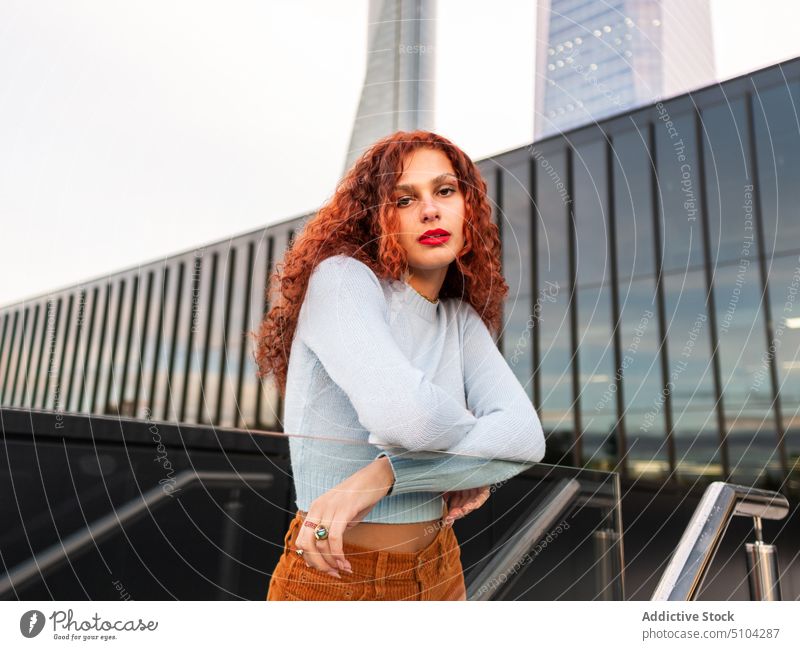 Self assured redhead woman leaning on glass fence on city street confident style district cool millennial serious portrait appearance charismatic female young