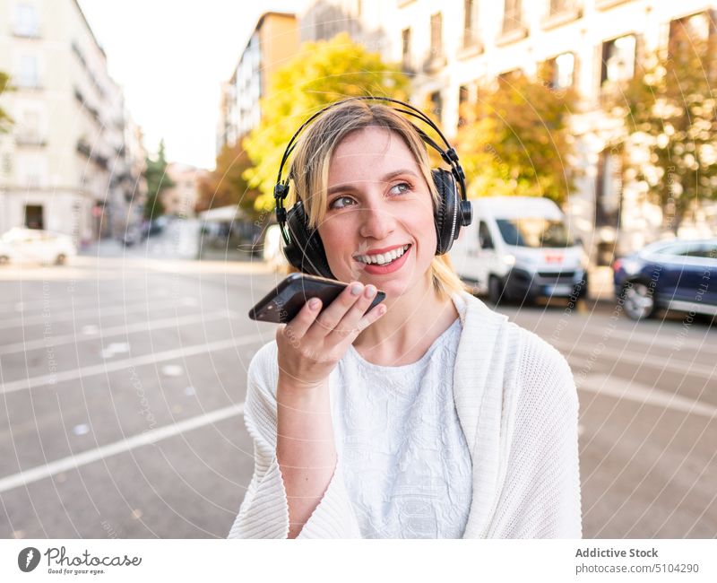Happy young lady in headphones using smartphone and smiling on street woman voice message record positive city music smile happy audio female blond casual