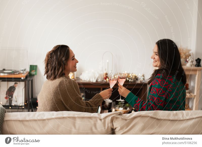 Two women toasting with sparkling wine glasses at party, drinking to celebrate a success or holiday. St Valentines or Galentine's Day. Friends celebrating holiday season sitting on a couch at home apartment and drinking, being happy, smiling and laughing.