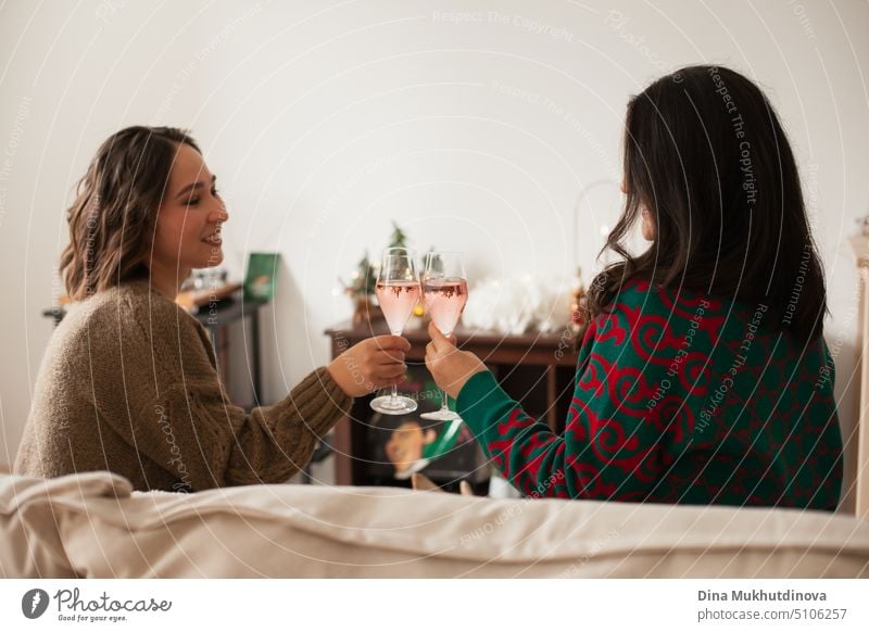 Two women toasting with sparkling wine glasses at party, drinking to celebrate a success or holiday. St Valentines or Galentine's Day. Friends celebrating holiday season sitting on a couch at home apartment and drinking, being happy, smiling and laughing.