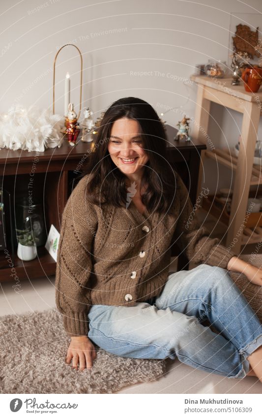 Candid photo of a woman jeans and cardigan sitting at home decorated for Christmas holidays, celebrating. Smiling woman with brown hair, laughing. cheerful