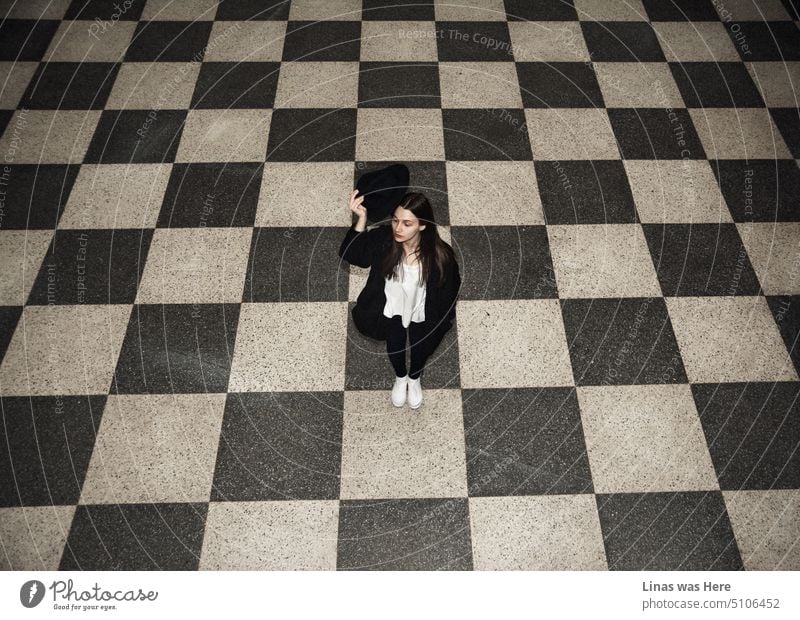 Minimalism is all over this image. From light & dark tiles to a gorgeous brunette girl. Dressed in a white shirt and a black outfit in general. While wearing white sneakers. A pretty woman is being all fashionable and cute.