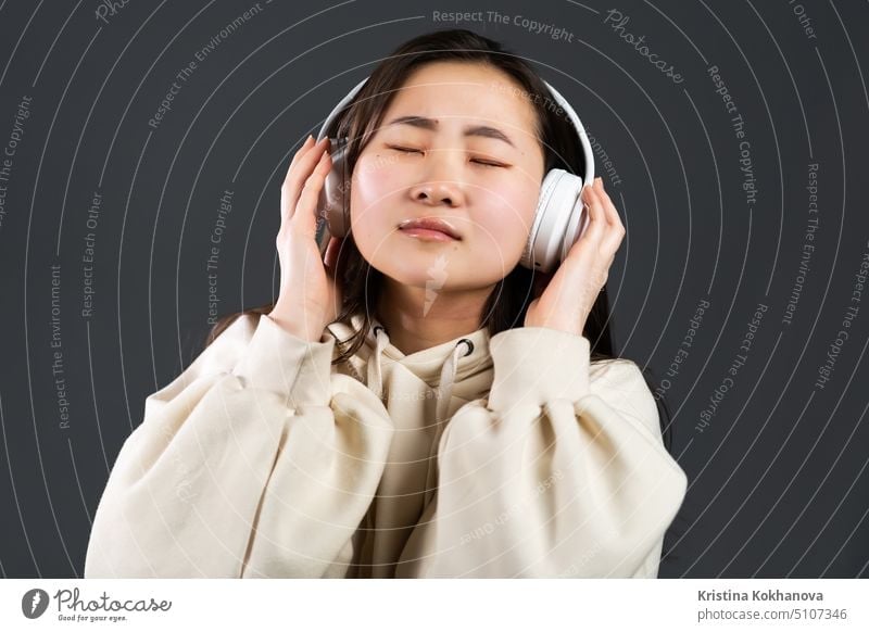 Young beautiful asian woman with headphones in modern wear enjoying and dancing on grey background. Radio, melody, technology concept. light glamour fashion