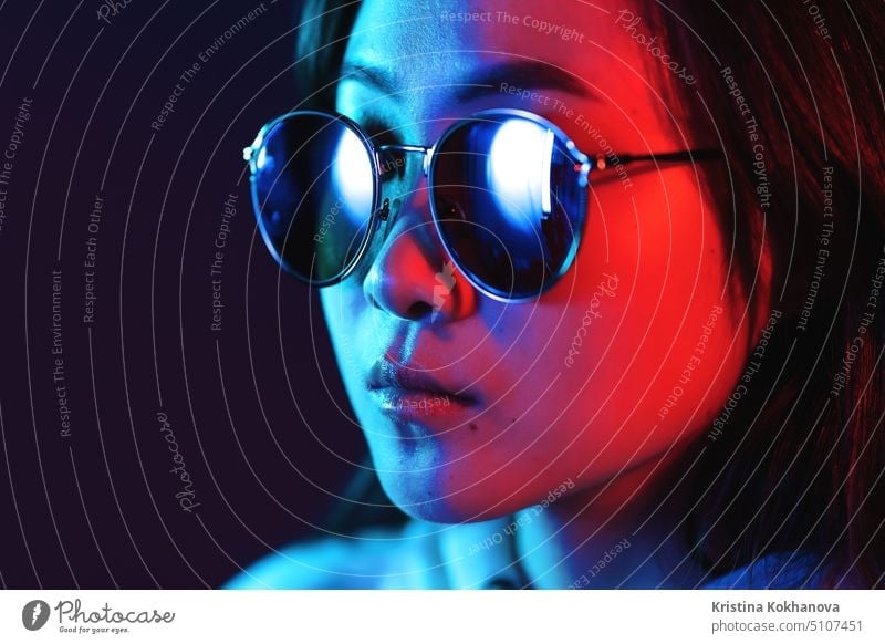 Young pretty girl with asian appearance in neon pink and blue lights at night. Serious trendy hipster teenager in glasses glamour fashion woman beauty model art