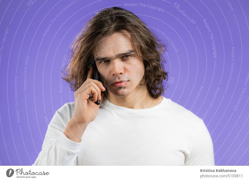 Young serious man speaks on phone. Guy is angry, holding and using smart phone. Violet studio background. person mobile phone communication technology adult