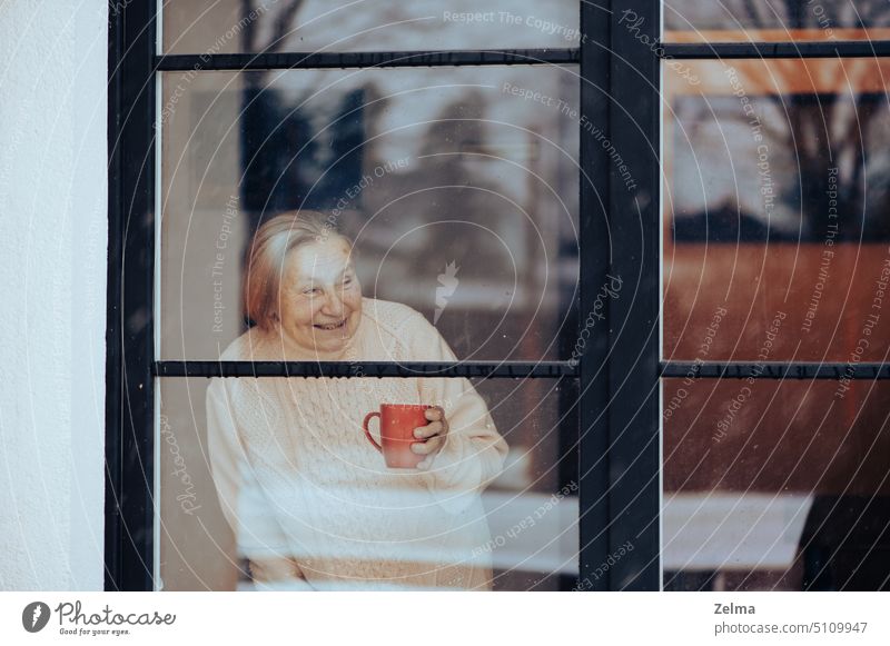 smiling old woman with a cup of coffee looks through a dirty window glass senior elderly female looking smile face nursing home house indoors loneliness sad