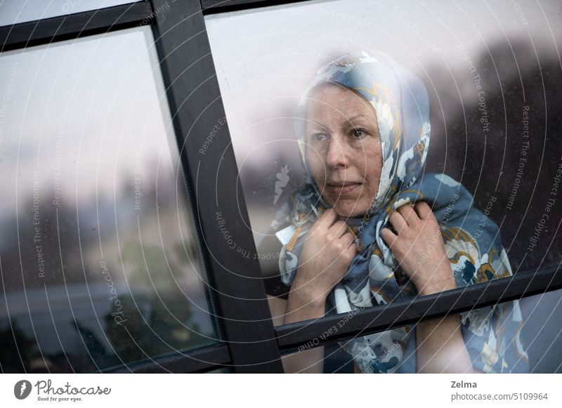Smiling middle-aged woman with scarf on head looking through dirty window glass mature elderly behind face expression hands indoor old senior disease ilness