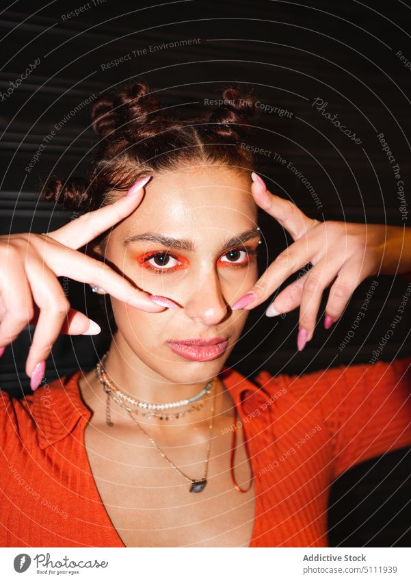 Stylish woman touching face and looking at camera serious trendy makeup eyeshadow orange style accessory portrait bantu knots fashion appearance model wall