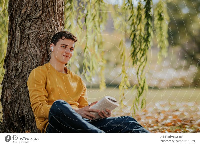 Happy man sitting under tree reading book using earphones park nature willow autumn gadget male device smile happy foliage listen leaf lifestyle entertain