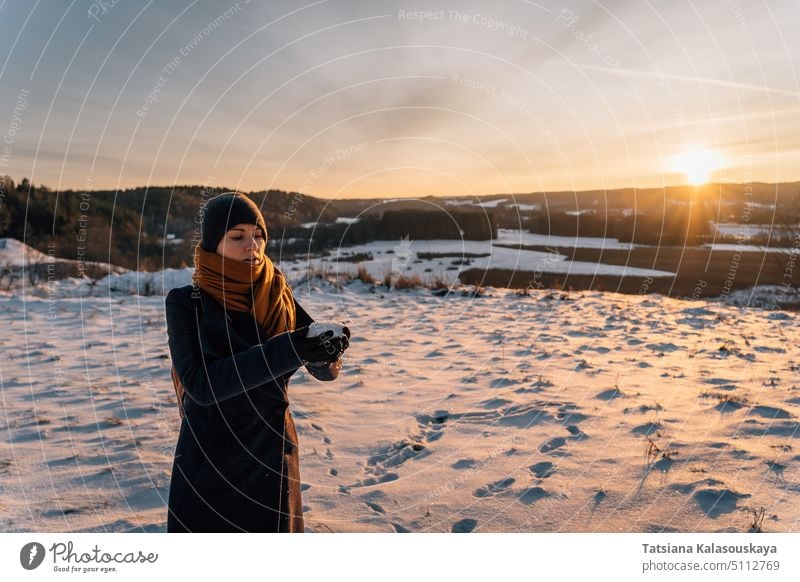 A young woman in a coat and hat at sunset stands on a snow-covered field and holds a handful of snow in her hands hand. winter warm clothes female people person