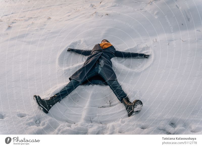 A young woman makes a snow angel lying in the snow with her arms and legs spread to the sides winter warm clothes lie female make snow people person European