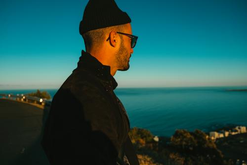 Man with sunglasses and cap looks at the sea from a viewpoint Cap Sunglasses Ocean vantage point Human being Blue laterally side view Meditative adult Hipster