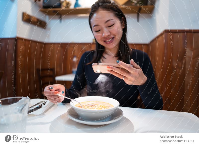 Cheerful young Asian woman taking photo of soup in restaurant take photo smile smartphone dinner delicious ramen food delight mobile female asian ethnic