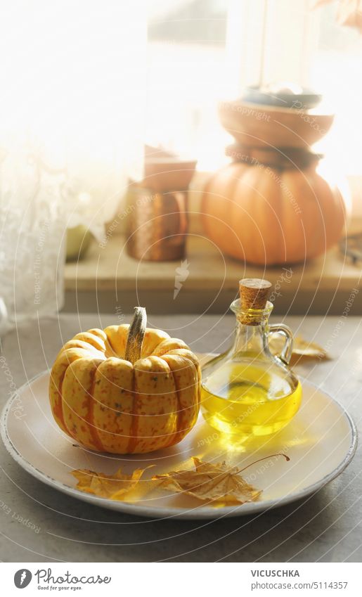 Little pumpkin, cooking oil in glass decanter and yellow autumn leaf on plate at window background with natural light. Cooking at home with seasonal fall vegetables. Front view.