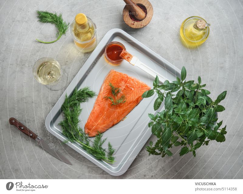 Preparation of salmon fish fillet on baking tray with healthy ingredients: herbs, spices, wine, oil and kitchen utensils on table. Healthy cooking with fresh fish at home. Top view.