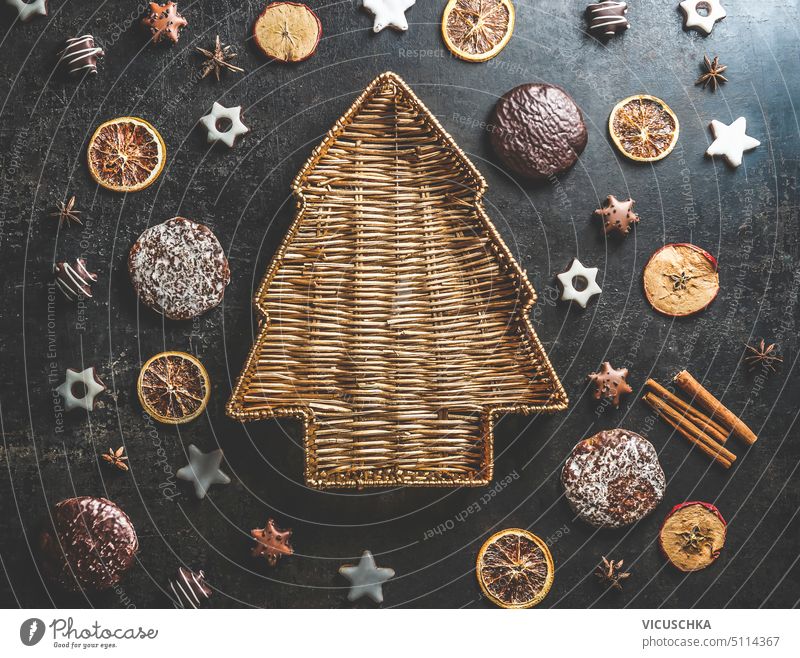 Christmas tree shaped wooden tray on dark concrete table with gingerbread, cookies, dried orange slices and cinnamon sticks. Delicious seasonal winter sweets and flavors. Top view.
