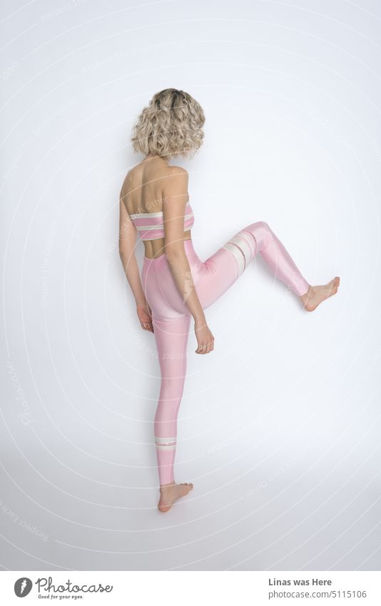 A minimalistic shoot in a studio. A model test of a curly blonde-haired girl. Just kicking a wall in her pink shiny sports outfit. Sports fashion with some action.