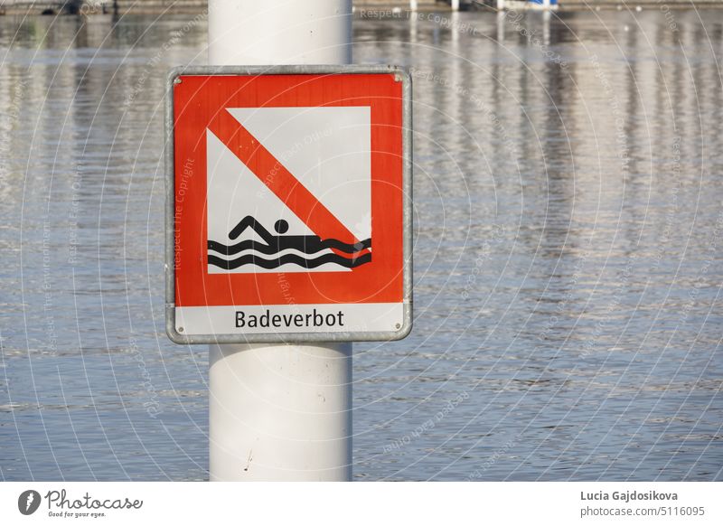 Red square signpost saying in German language bathing prohibited attached on metal pole. Placed in Lake Lucerne in Switzerland. The lake itself is on background. It is warning symbol against danger.