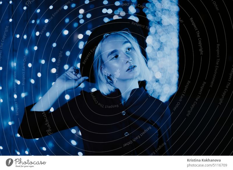 Millennial enigmatic pretty girl blond hairstyle near blue glowing neon wall at night. Hair, hipster hat, nose piercing. Mysterious woman people adult city