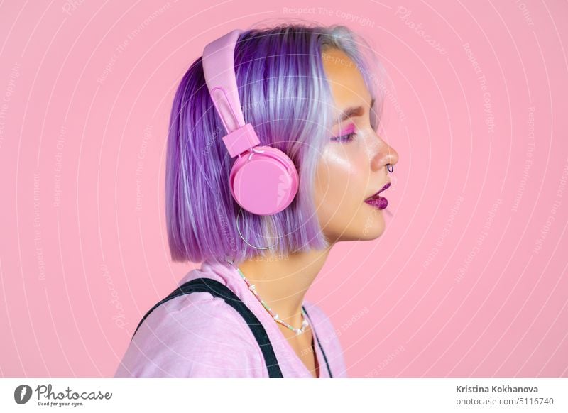 Close-up portrait of pretty girl with dyed violet hair listening to music, smiling, dancing in headphones in studio against pink background. Music, dance, radio concept