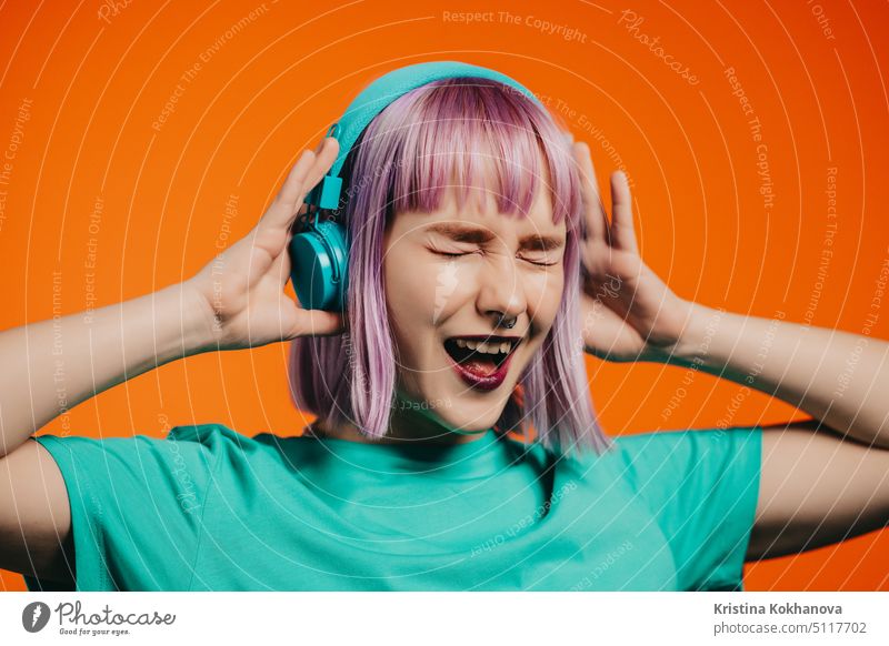Exceptional woman with dyed violet hair listening music in headphones and singing on orange background. Unique hipster girl dancing person female fun studio