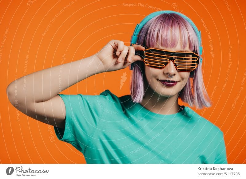 Exceptional woman with dyed violet hair listening music in headphones and singing on orange background. Unique hipster girl dancing with lattice sunglasses.