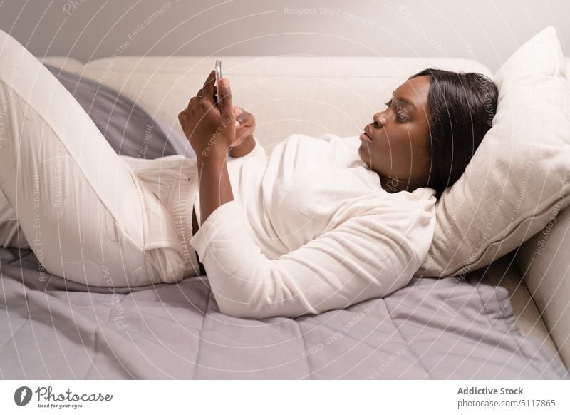 African American woman using smartphone on sofa social media pajama rest home evening addict female young black african american blanket pillow living room