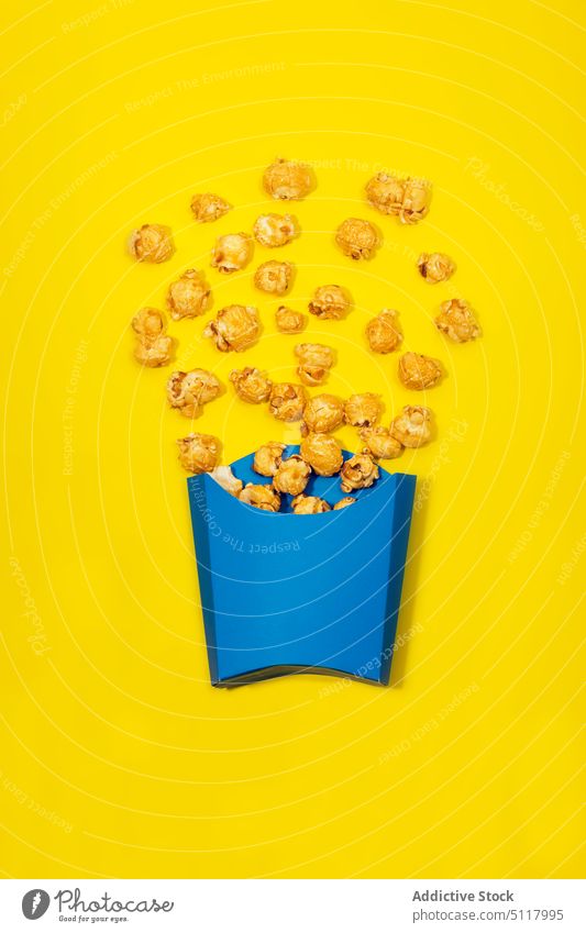 Paper bag with caramel corns scattered on yellow surface popcorn sweet snack dessert delicious yummy tasty calorie unhealthy appetizing treat bright food