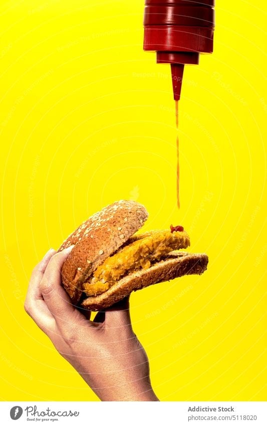 Crop woman pouring tomato sauce on burger in yellow studio ketchup fast food unhealthy homemade junk food hungry tasty patty bun female hand appetizing yummy
