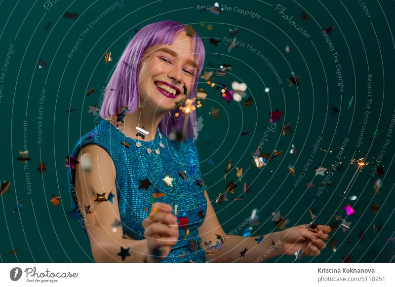 Hipster woman with sparkling bengal fire dancing under confetti rain on green background. Christmas holiday concept. Young dyed violet haired girl with sparkler celebrating, smiling, enjoying time.