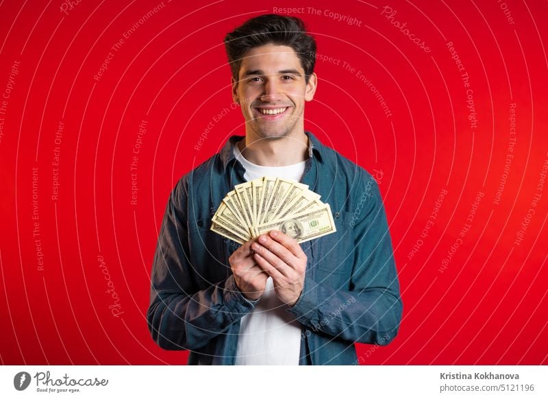 Satisfied happy excited man showing money - U.S. currency dollars banknotes on red wall. Symbol of success, gain, victory. rich cash finance bill male person